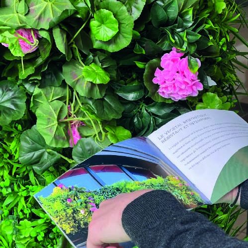 Main d'un homme tenant un livre ouvert devant un mur de plantes vertes et fleurs roses.