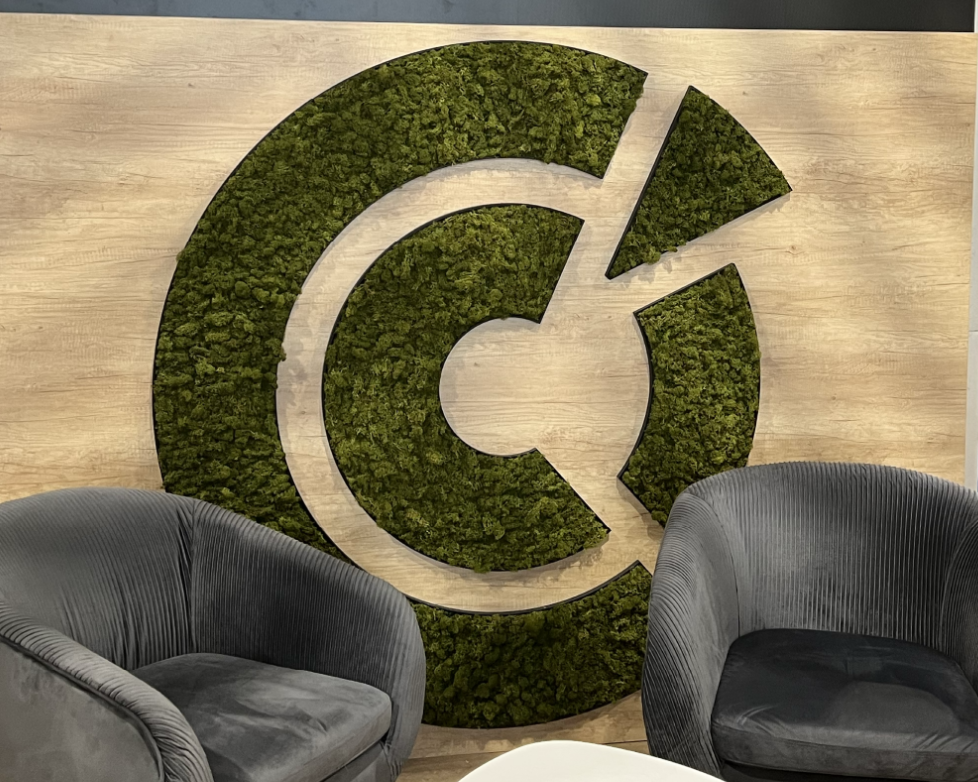 Logo en mousse verte sur fond en bois, avec des sièges gris en avant-plan.
