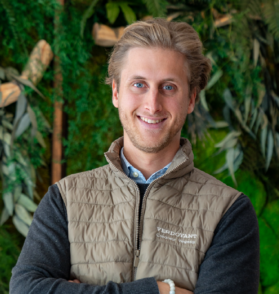 Homme souriant avec un gilet beige, bras croisés, devant un mur végétal.